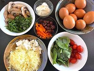 Bowls of fresh ingredients for frittata, including eggs, cheeses, spinach, tomatoes, mushrooms, peppers, olives, and herbs.