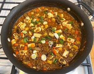 Hearty lentil and tofu stew simmering in a skillet with carrots, sweet potato, mushrooms, and green onions