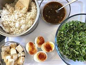 Ingredients for poke bowl: pot of steamed rice, bowl of microgreens, soy-ginger dressing, cubed tofu, and hard-boiled eggs sprinkled with paprika.