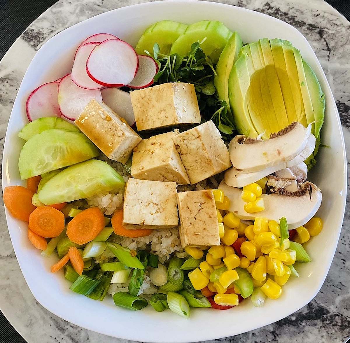 Close-up of a vegetarian poke bowl featuring tofu cubes, avocado, cucumbers, radishes, mushrooms, corn, scallions, carrots, and fresh microgreens over rice.