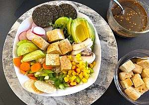 Serving of a poke bowl with tofu, avocado, mushrooms, radish, cucumber, corn, and carrots, topped with scallions and microgreens; soy-ginger dressing and marinated tofu cubes on the side.