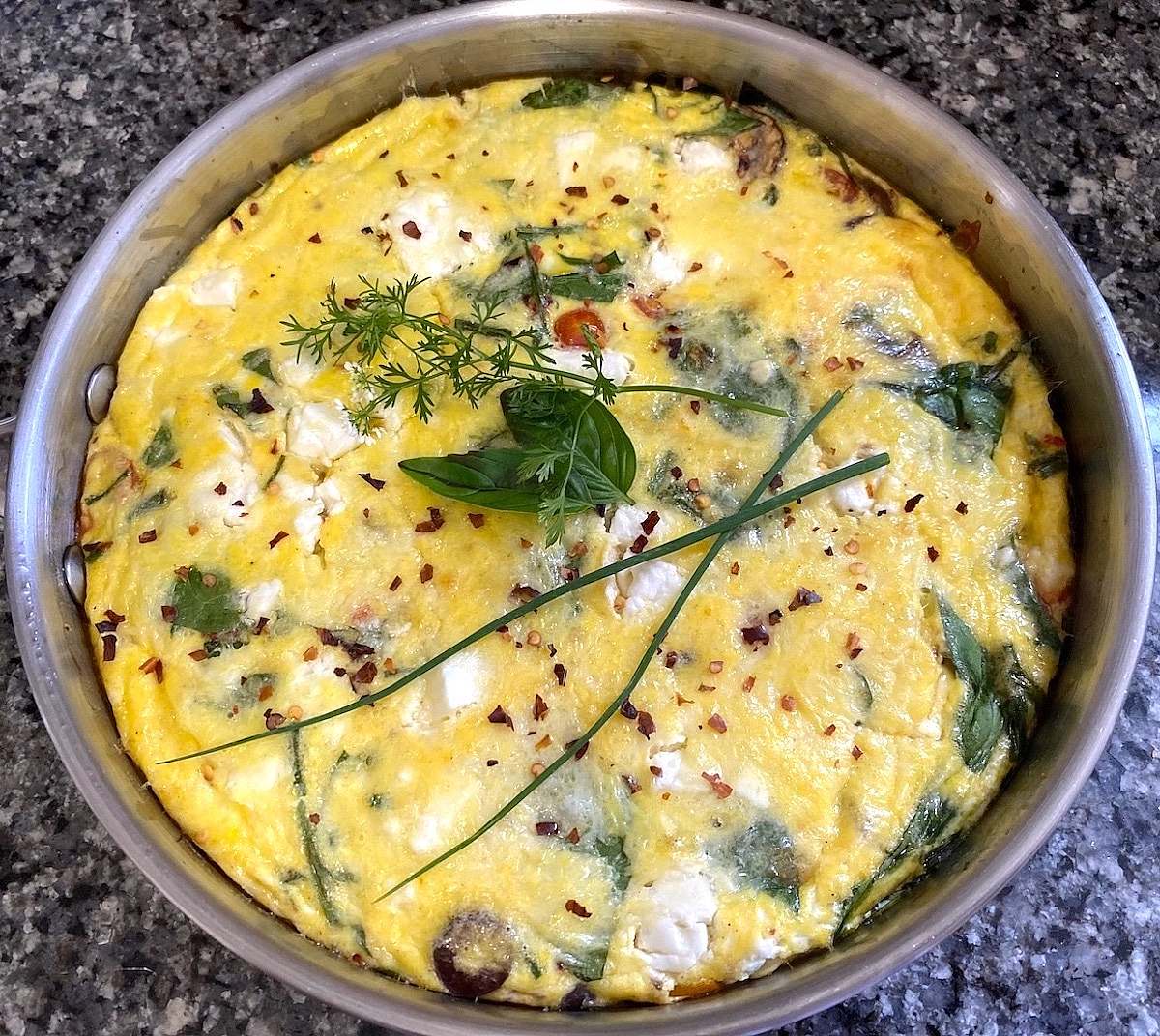 Freshly baked Mediterranean frittata in a stainless steel skillet, topped with herbs and red pepper flakes.