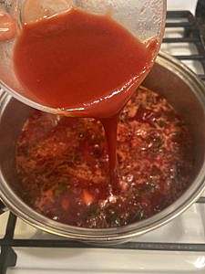 Tomato purée being poured into a pot of simmering vegetable borscht.