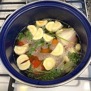 A blue pot filled with simmering clear fish broth base with carrots, parsnip, onion, tomatoes, parsley, and garlic.