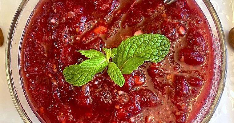 Homemade cranberry sauce with chopped almonds, candied ginger, and fresh mint garnish in a glass bowl. The sauce has a deep red hue and glossy texture, surrounded by whole almonds and mint leaves.