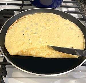 Thin crêpe cooking in a skillet, edges curling slightly as it’s flipped with a knife.