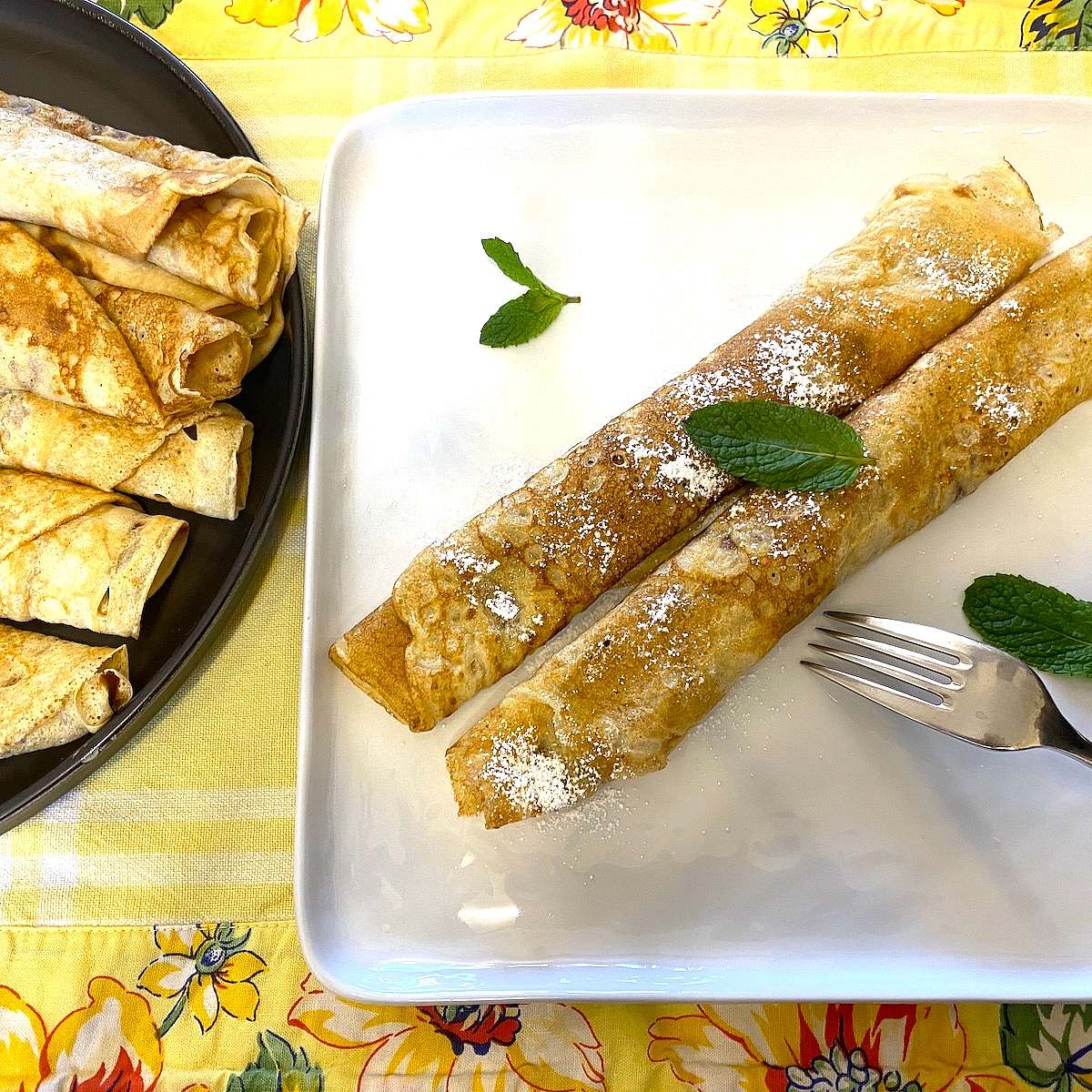 Two rolled crêpes on a white plate, dusted with powdered sugar and garnished with mint leaves.