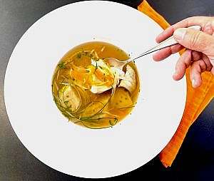 A bowl of clear fish soup garnished with julienned vegetables, herbs, and olive oil, served with a spoon and an orange napkin.