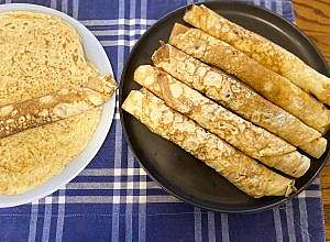 Black round plate with several rolled crepes next to one rolled crepe on white plate.
