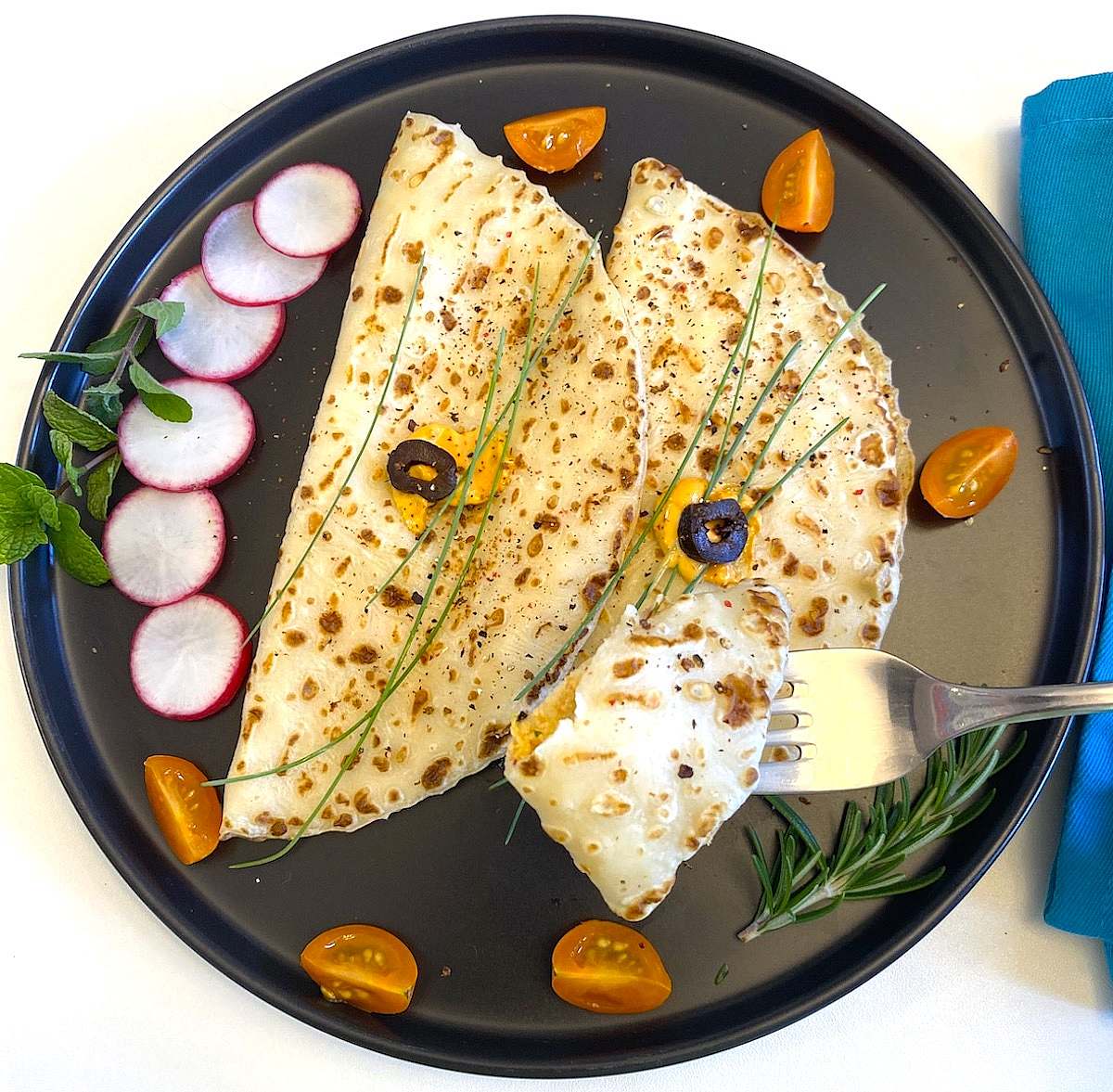 Two savory crêpes on a black plate with herbs and tomatoes, with a fork lifting a small piece.