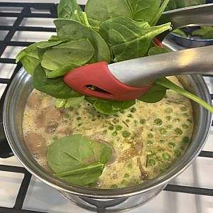 Fresh baby spinach being added with tongs to a simmering pot of vegetable curry soup.